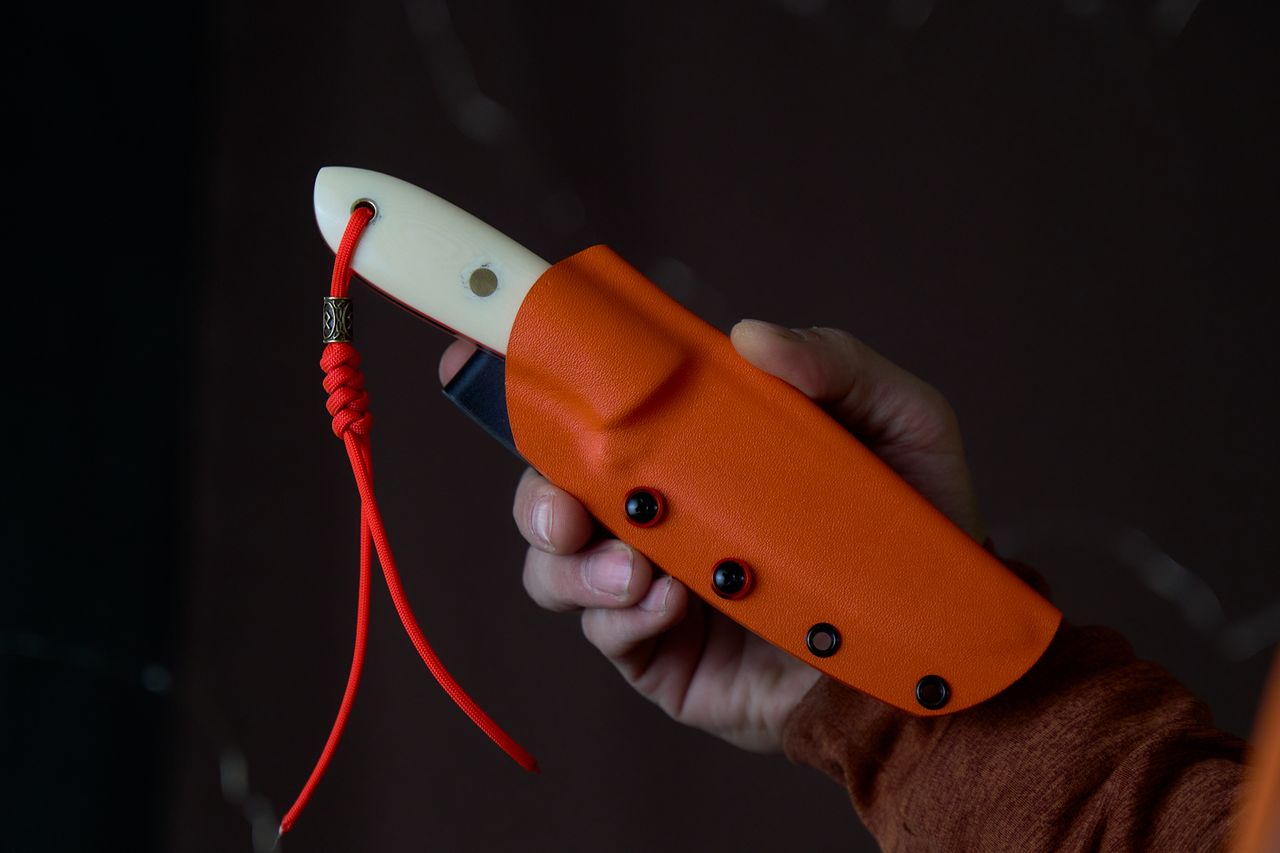 Close-up of a hand holding a knife in a vibrant orange sheath with a red cord.